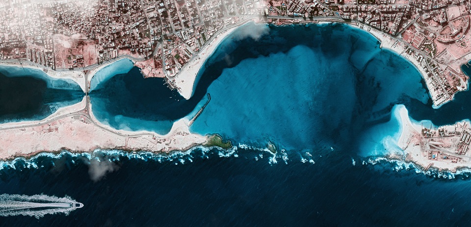 Aerial image of a coastline with a city
			and a boat with a wake sailing by